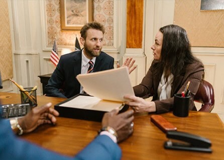 A man and woman in business attire sit at a desk discussing papers. An American flag is visible in the background.
