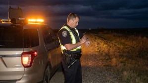 uniformed police officer writing notes beside patrol vehicle with flashing lights at night